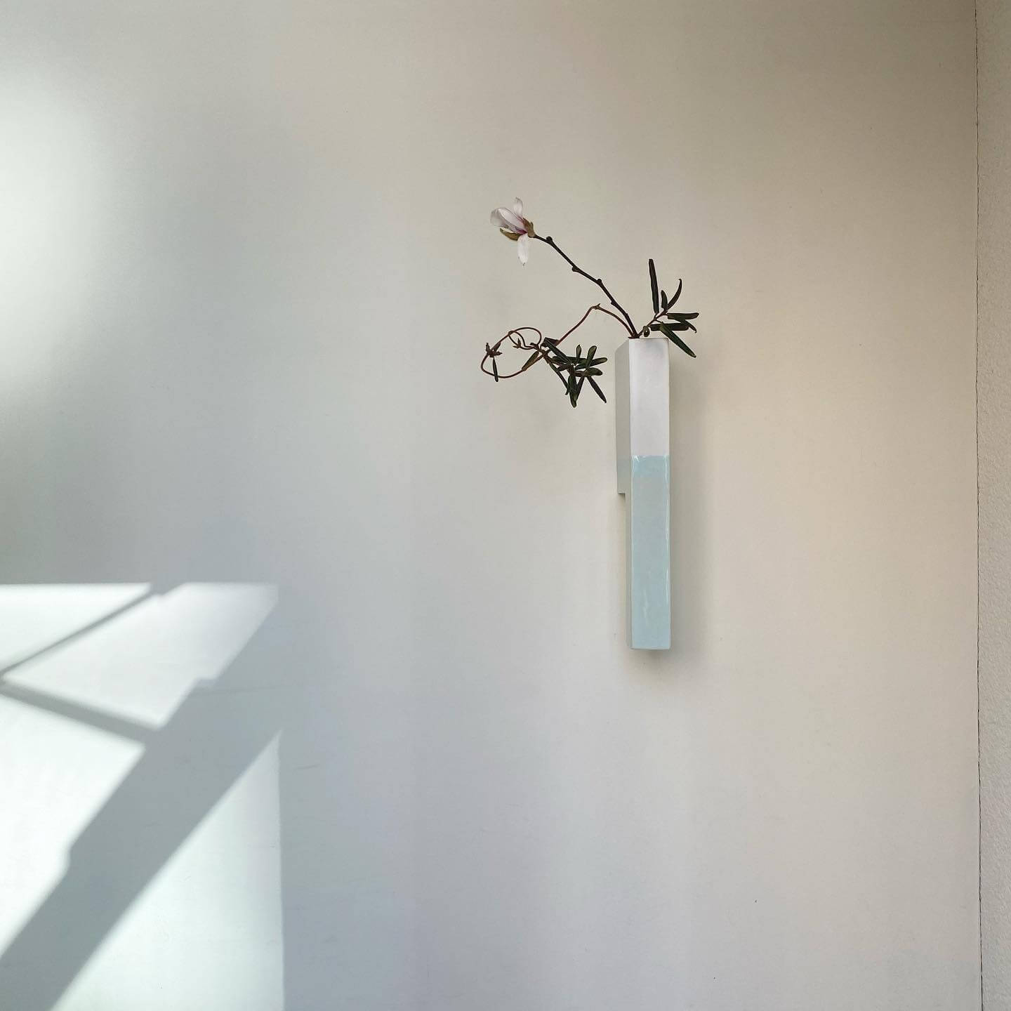 写真：茶室の壁に飾るような、壁掛け型の生け花。白い細長い四角柱型の器に白いこぶしのような花が一輪生けられている
