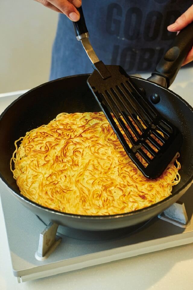 写真：フライパンに麺を広げて、ぎゅうぎゅう押しつけながら焼く様子。麺にきれいな焦げ目がついている