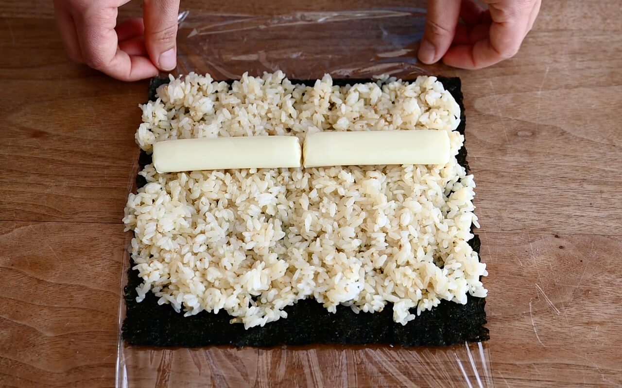 ラップの上に海苔を置き、上側を1cm残してご飯を広げる。 真ん中より下あたりに、さけるチーズを横長に並べ、手前のラップを持ち上げ、チーズを芯にして、軽く握るように巻く。