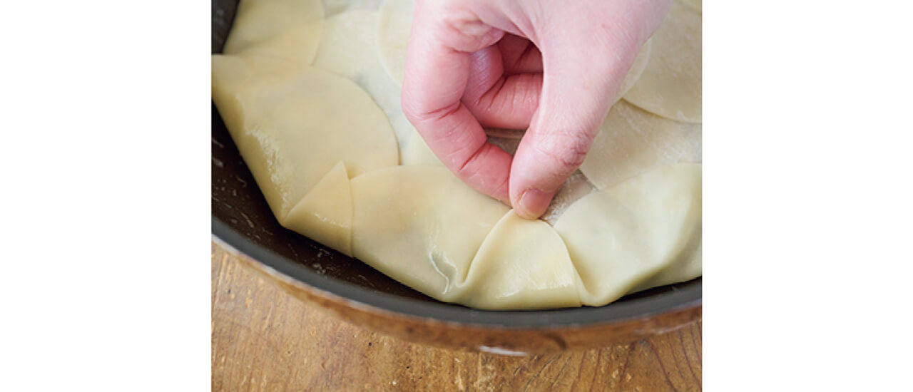 フライパンの端の餃子の皮に水をつけ、ひだをとりながら折って包む