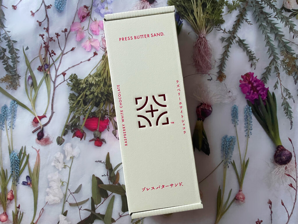生成りがかった白い箱に赤いロゴが入った、PRESS BUTTER SANDの「ラズベリーホワイトショコラ」パッケージ。背景には球根植物をメインとした美しい草花の写真が敷かれている。