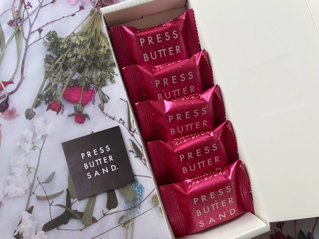 PRESS BUTTER SANDのラズベリーホワイトショコラ。白い箱にメタリックピンクの個包装が整然と並ぶ。背景にはグレーとグリーン球根植物を基調とした花柄とショップカード。