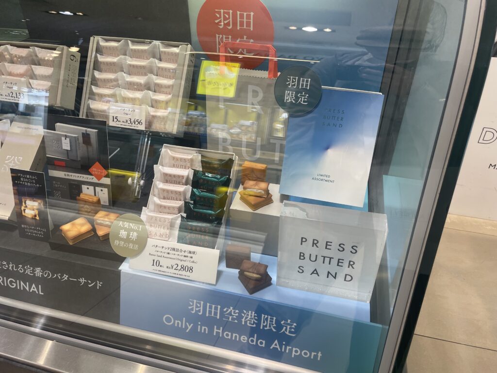 羽田空港限定プレスバターサンドのショーケース展示。人気商品「限定アソート」や「珈琲×キャラメル」などの詰め合わせが並ぶ、Haneda Airportでしか買えない限定土産。