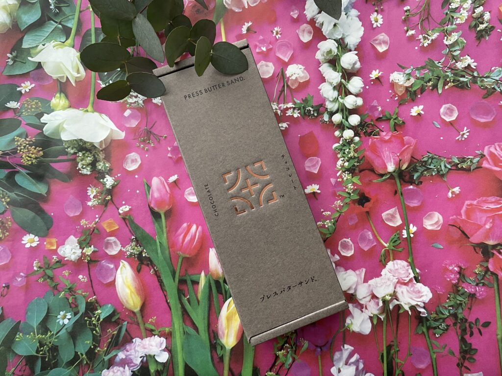 グレーブラウンの箱に銅色のロゴが施された、PRESS BUTTER SANDの「チョコレート」フレーバーのパッケージ。背景はピンク色の花々と結晶のような装飾が散りばめられた華やかな写真。
