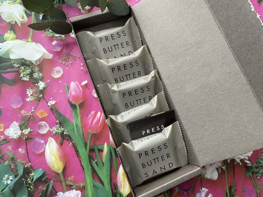 PRESS BUTTER SANDのチョコレート味。グレーの箱にグレーベージュの個包装が並ぶ。背景はピンクや白のチューリップをあしらった華やかなデザイン。