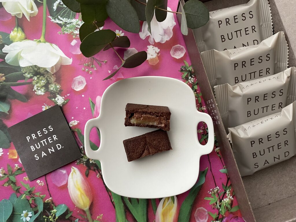 ピンクの花柄の背景に、白い皿に乗せたPRESS BUTTER SANDのラズベリーホワイトショコラと、個包装された同商品が並ぶ美しい構図の写真