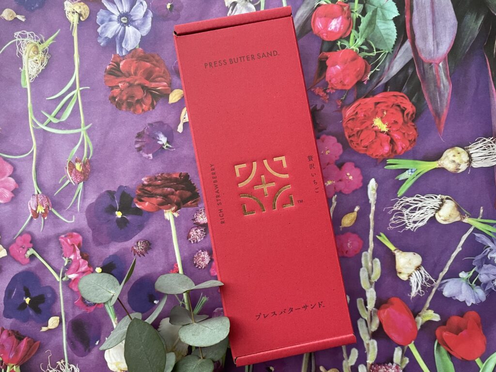赤い箱に金のロゴがあしらわれた、PRESS BUTTER SANDの「贅沢いちご」フレーバーのパッケージ。背景には紫系の花々の写真が敷かれている。