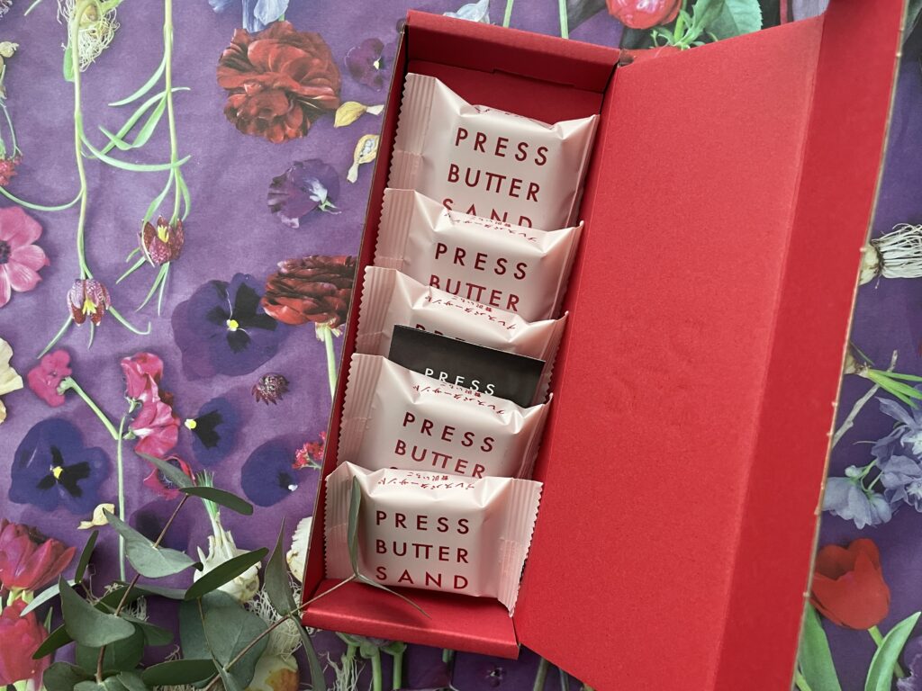 PRESS BUTTER SANDの贅沢いちご。赤い箱に淡いピンクの個包装が並ぶ。背景には紫を基調とした花柄のデザインとユーカリの葉。