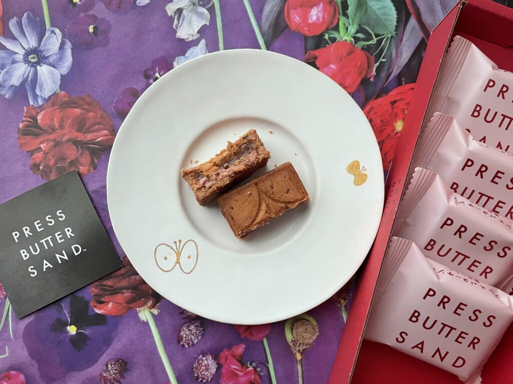 赤や紫の花柄の背景に、バターサンド〈贅沢いちご〉を載せた白い皿とピンクの個包装が映えるPRESS BUTTER SANDの季節限定商品の写真