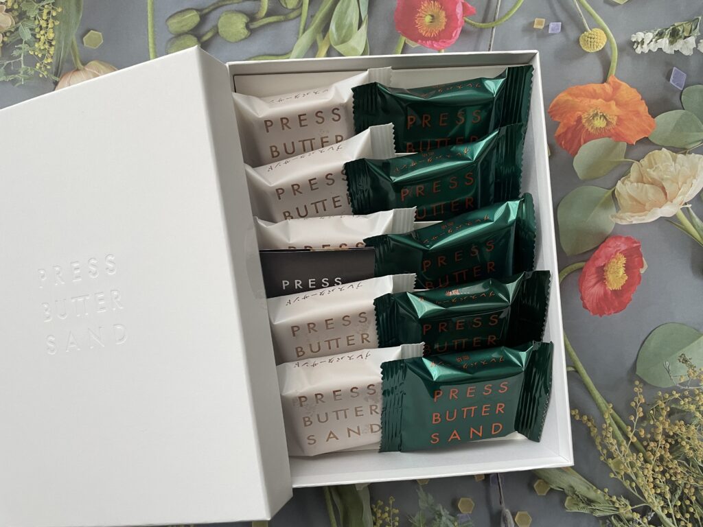 PRESS BUTTER SANDの羽田空港限定パッケージ。白い箱に白と緑の2種類の個包装が交互に並ぶ。背景には花柄のデザイン、黒いショップカードが添えられている。