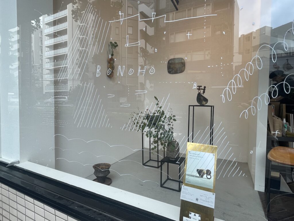 ガラス窓の外から見た、newtonでの佐藤尚理さんのオブジェ展示の様子。ガラスには佐藤尚理さんにより白いポスターカラーで手描きされた模様や「BONOHO」の文字も見える。