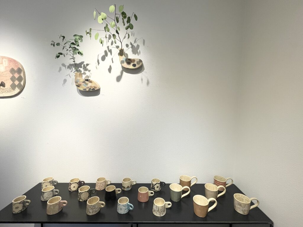 壁に飾られた植物入りの鳥形陶器花器と、テーブルに並ぶ多彩なマグカップの展示。シンプルな白壁と黒いテーブルが作品を引き立てている。