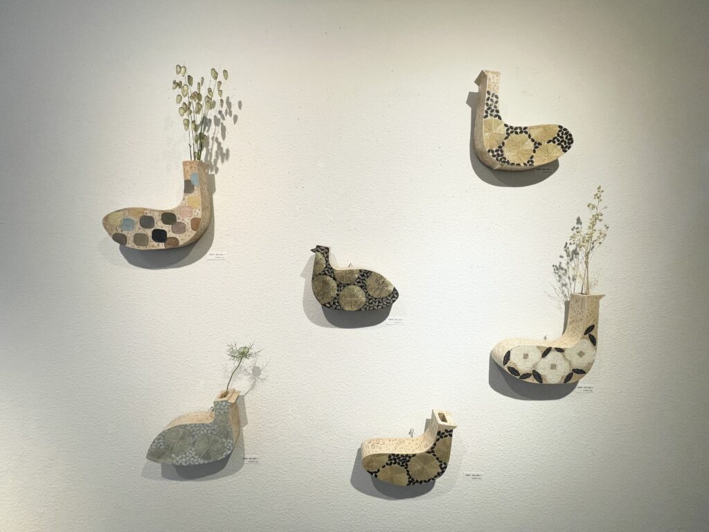 鳥の形をした陶器の壁掛け花器が白い壁に6点展示され、いくつかにはドライフラワーが挿されている。色とりどりの模様が施され、柔らかな雰囲気を持つ。