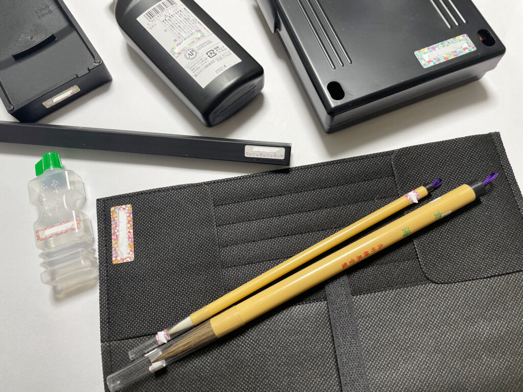 お習字セットの各道具に名前シールが貼られている。硯、墨汁、筆、文鎮、水差しなどが写っており、名前部分は加工で消されている。