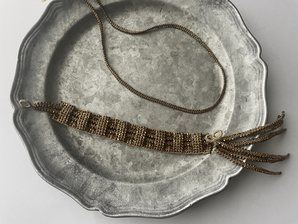 金属のトレイの上に置かれたゴールドのビーズブレスレット。繊細なビーズが織り込まれ、端にはタッセルがついている。