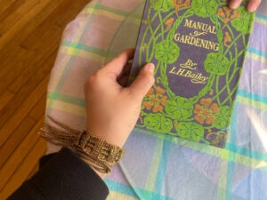 手首にゴールドのビーズブレスレットを巻いた手と、緑の洋書『MANUAL OF GARDENING』を持った様子。背景はパステルカラーのチェック柄クロス。