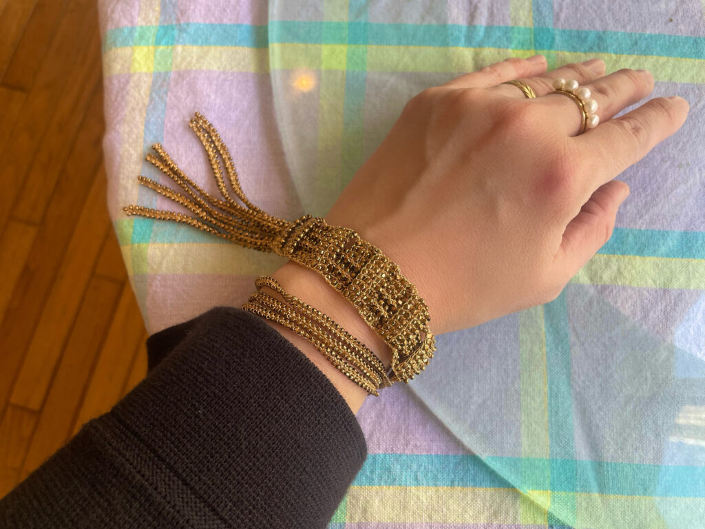 ゴールドのビーズブレスレットを着けた手で『MANUAL OF GARDENING』の表紙を持っている様子。リングを複数着用。背景はチェックの布。