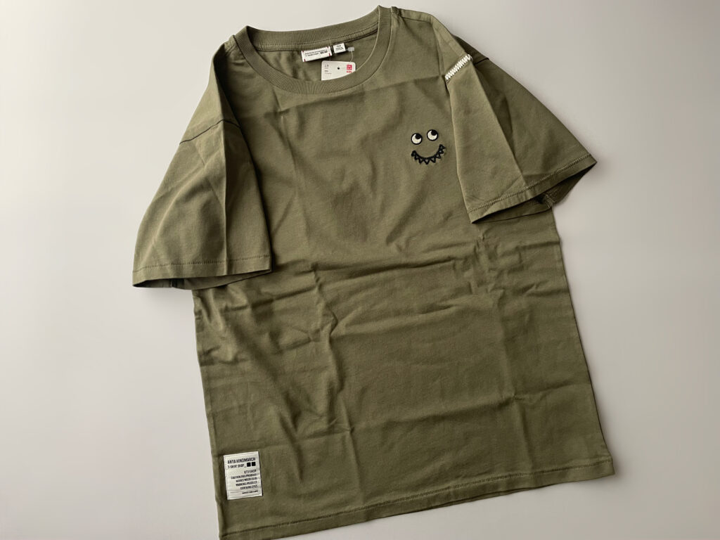 ユニクロ×アニヤ・ハインドマーチのコラボのTシャツ、KT1EASY/クルーネックTのGREEN、150cmサイズのフラットレイ画像。胸元にはEyesと口がギザギザの刺繍、左肩にはハンドステッチ風のジグザグのステッチが入っている。右前裾にはデザインラベルが貼付してある。