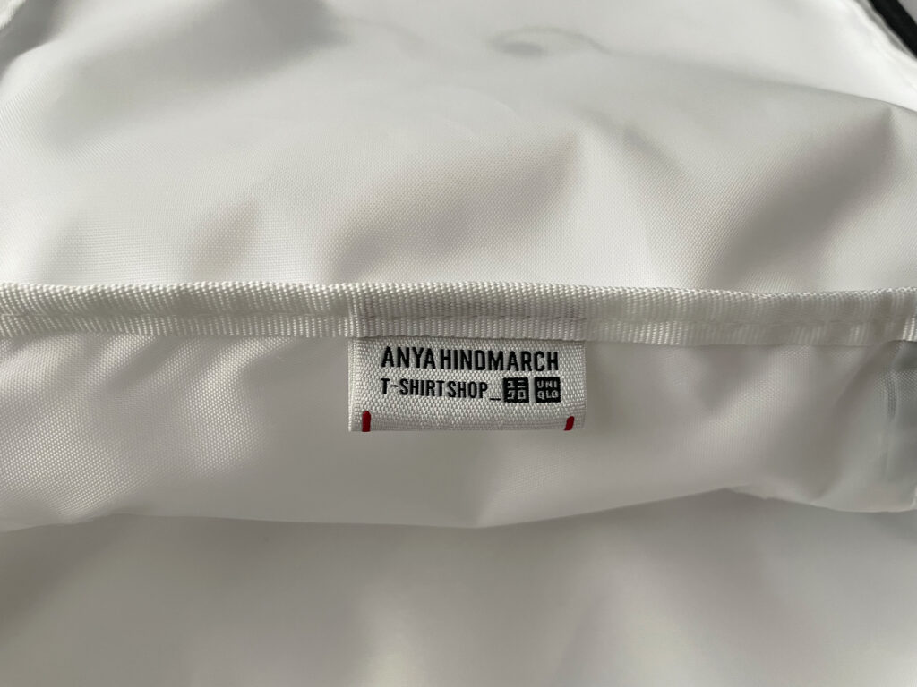 ユニクロ×アニヤ・ハインドマーチのコラボ2025のスクエアポーチの内側に付いているタグのアップ写真。ユニクロとアニヤ・ハインドマーチのダブルネーム入りで、“T-SHIRT SHOP”の文字が入っている。