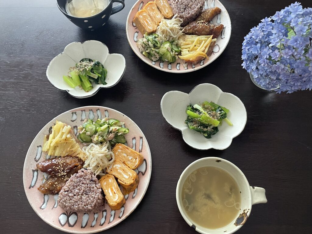 セブンイレブン×結わえるコラボの「寝かせ玄米®おむすび」がのったワンプレートランチの写真。アジの南蛮漬けや卵焼き、牛肉とじゃがいもの重ね焼きや、きゅうりの和え物、モヤシのナムルなどがのっている。サイドには小松菜の塩お浸しやお味噌汁も並ぶ。傍らには紫陽花が活けてある。