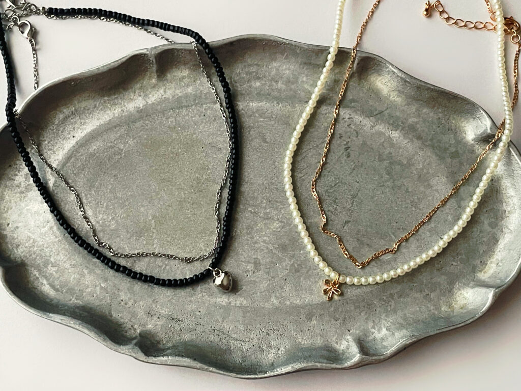 GUの390円ネックレス2種類。左は黒ビーズとハートモチーフのネックレス、→はパールとリボンモチーフのネックレス。寄りの写真。下にはシルバーのレリーフプレートが敷かれている。