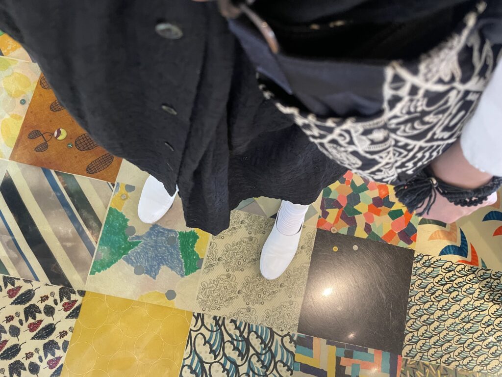 40代女性の2025年7月中旬コーデ、Sa-Rahの黒の総レースワンピースとqueの白のplainシューズを合わせている。床はミナペルホネンのテキスタイル柄がパッチワークのように敷き詰められている。