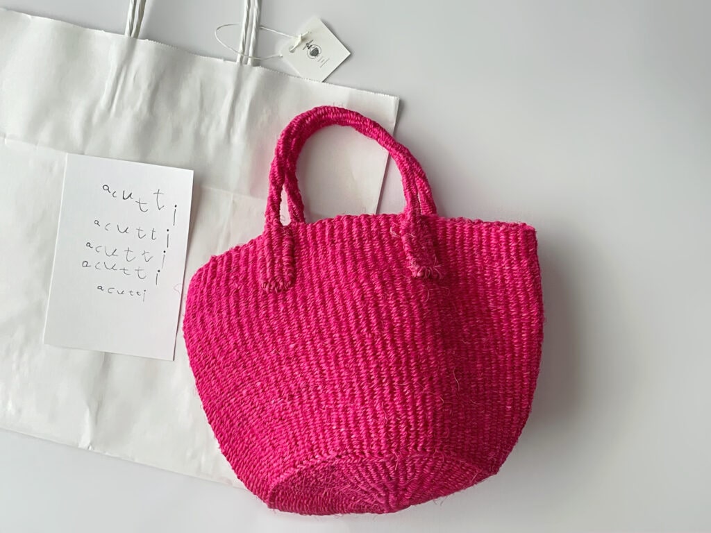 ケニア製のローズピンク色のザイザルかごバッグ。acuttiのショップカードが隣に並んでいる。