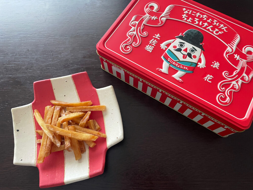 なにわちょろけんのちょろけんぴ缶と、本郷里奈さん作陶の、赤と白のストライプの十字型のお皿に芋けんぴがのった写真。