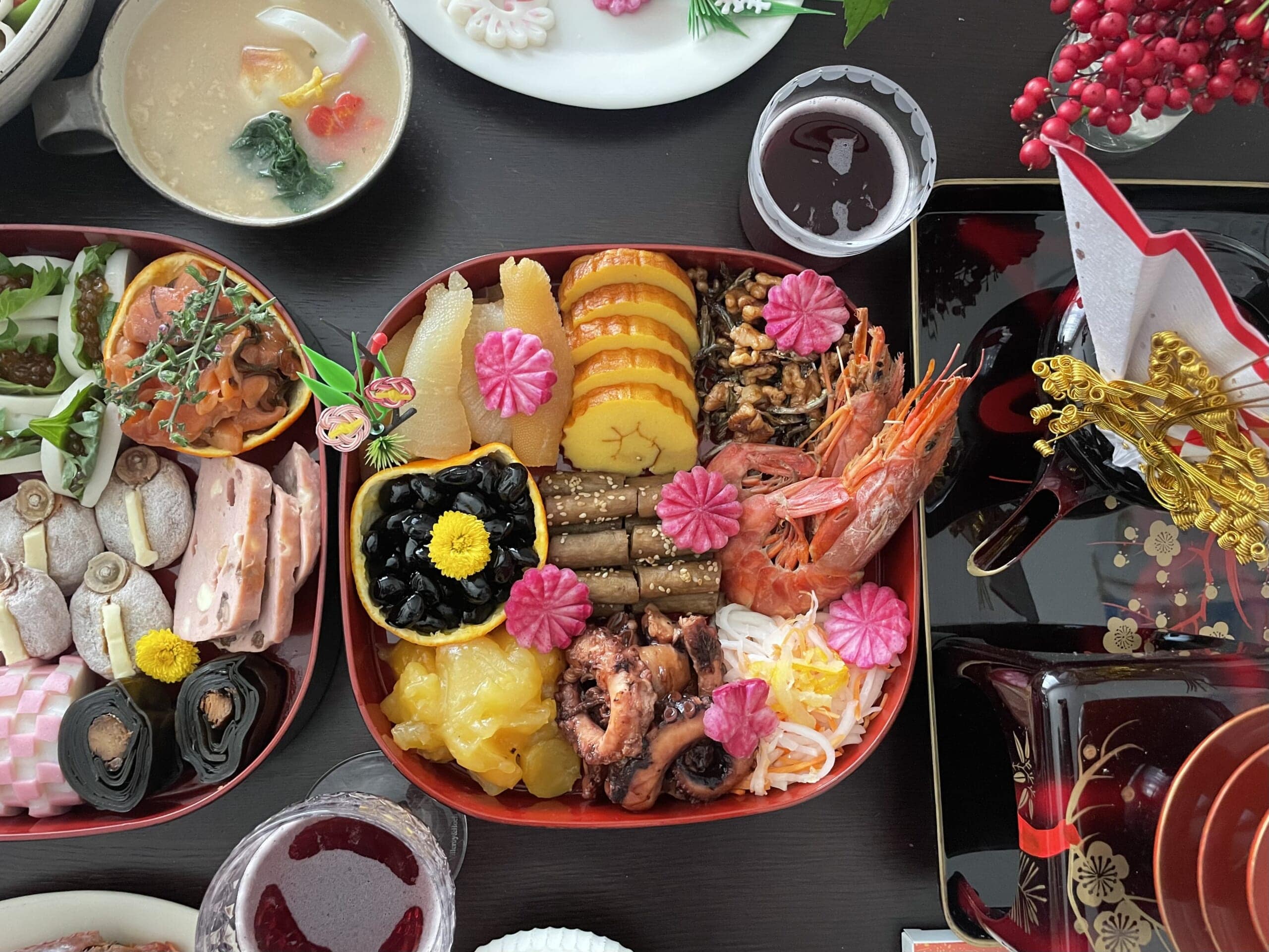 おせち2026の一の重。数の子、伊達巻き、くるみ田作り、黒豆、たたきごぼう、エビ煮、栗きんとん、タコの柔らか煮、大根とにんじんの生酢が詰められている。菊型に飾り切りされた赤大根がちりばめられている。