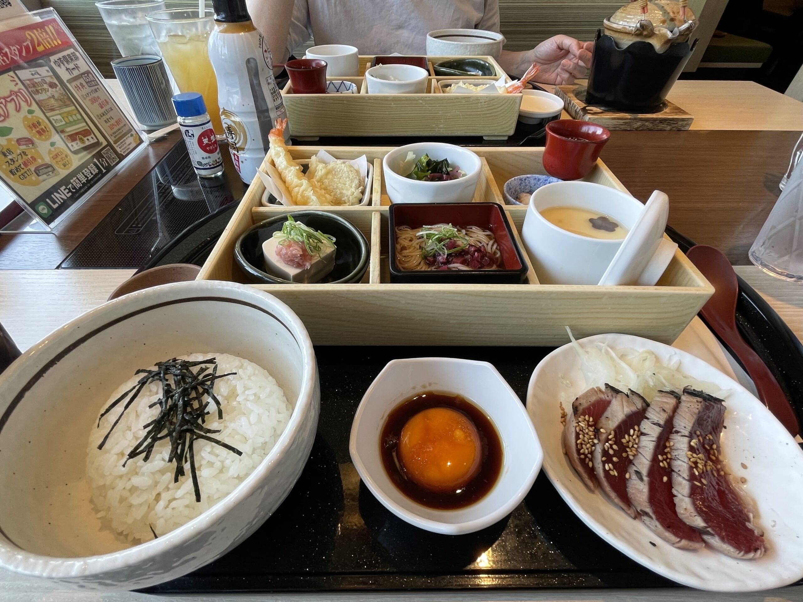 2026年4月の松花堂ランチ。茶碗蒸しや蕎麦、天麩羅、マグロ豆腐、もずく酢、サラダ、カツオのたたき、塩昆布茶漬けなどが並ぶ。